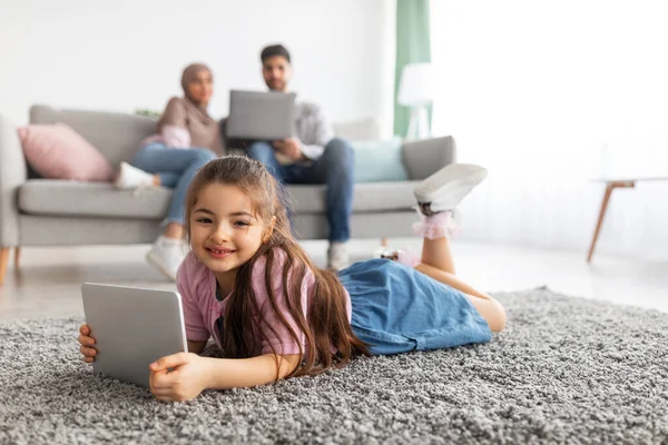 Söt östra flicka med hjälp av tablett, ligger på golvet matta och ler mot kameran, föräldrar avkopplande på soffan med laptop — Stockfoto