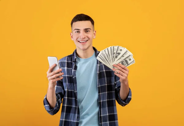Concepto de finanzas online. Positivo millennial hombre celular y dólares americanos sobre fondo naranja —  Fotos de Stock