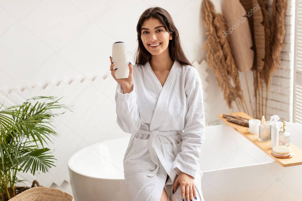 Woman In Bathrobe Showing Shampoo Bottle Advertising Product In Bathroom