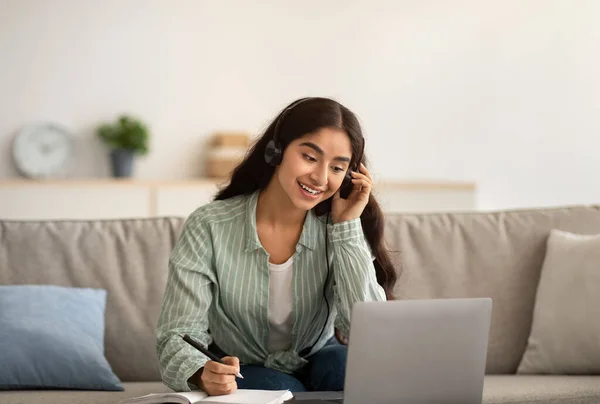 Gelukkig Indiase zakenvrouw in koptelefoon met online zakelijke bijeenkomst op laptop, het opschrijven van informatie thuis — Stockfoto