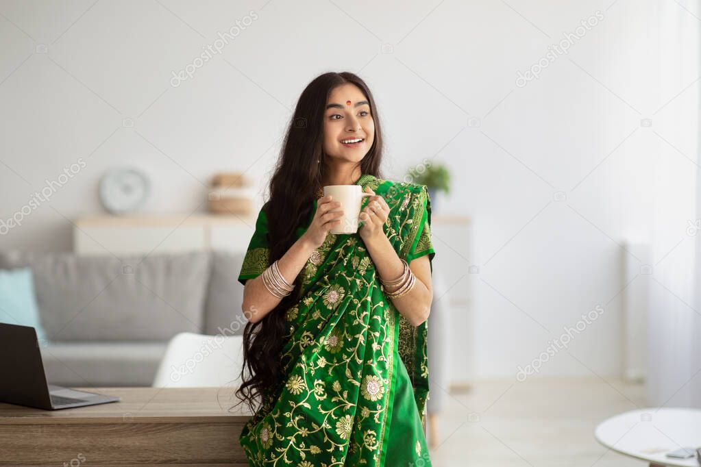 Pretty young Indian woman in green sari dress having coffee break at home