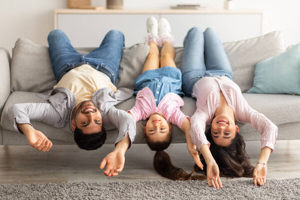 Upside down portrait of happy young family lying on sofa with heads down. Mother, father and daughter having fun