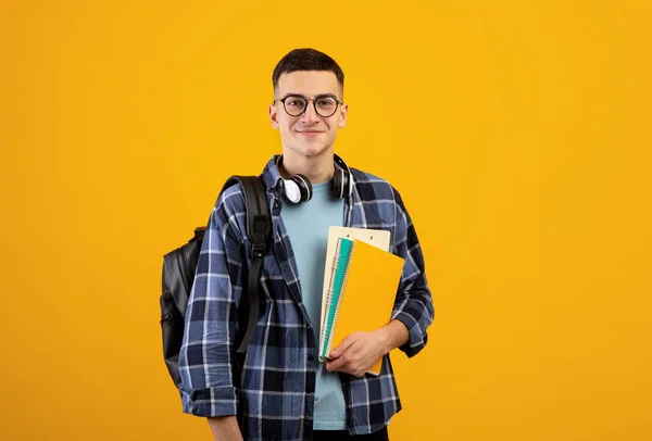 Onderwijsconcept. Positieve jongeman met rugzak, koptelefoon en notebooks poserend op oranje achtergrond — Stockfoto