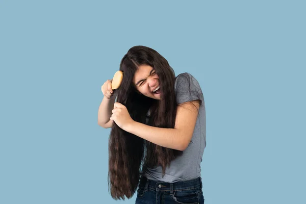 Frustrated Indian teen girl trying to brush her tangled long locks on blue studio background. Hairdressing and beauty concept — Stock Photo, Image