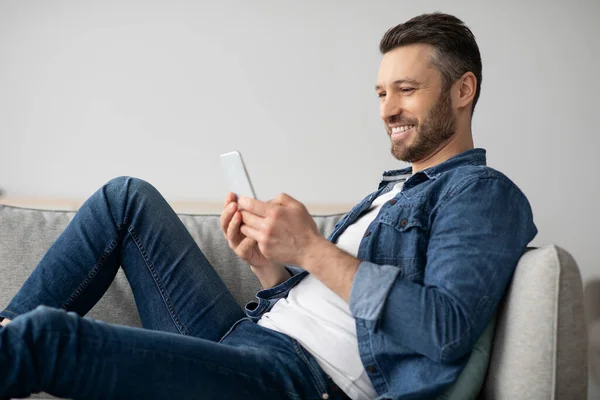 현대의 스마트폰을 사용하여 집에서 소파에 앉아 쉬고 있는 행복 한 남자 — 스톡 사진