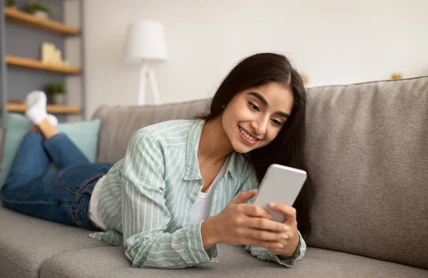 Retrato de milenar senhora indiana usando celular para trabalhar ou estudar, deitado no sofá em casa — Fotografia de Stock