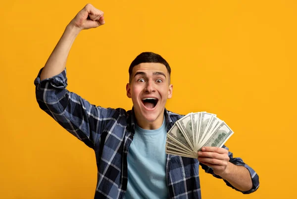 Chico hipster muy alegre sosteniendo un montón de dinero, gritando WOW y haciendo un gesto SI en fondo de estudio naranja —  Fotos de Stock