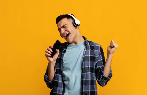 Emotionele jongeman in hoofdtelefoon die naar muziek luistert, liedjes zingt, smartphone gebruikt als microfoon op oranje achtergrond — Stockfoto
