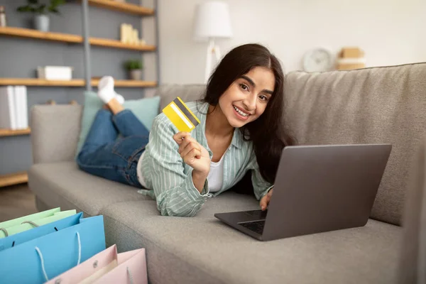 İnternetten alışveriş yapan güzel Hintli kadın, dizüstü bilgisayarı ve kredi kartıyla kanepede uzanıyor, hediye çantaları yakınlarda duruyor. — Stok fotoğraf
