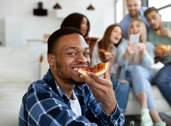 Aantrekkelijke zwarte man eten lekker pizza, zijn diverse vrienden met mobiele telefoon plezier op de achtergrond — Stockfoto