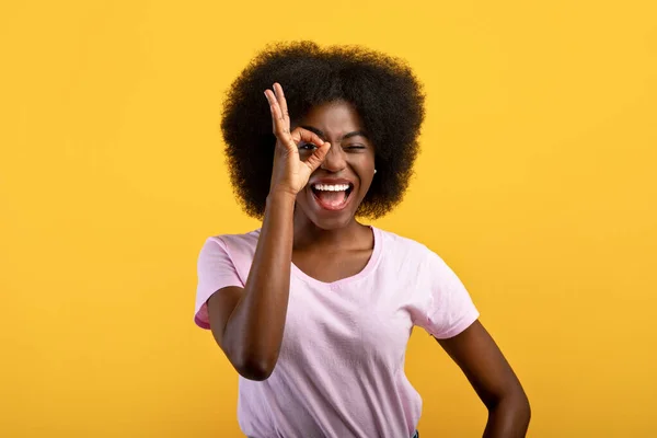 Her şey yolunda. Pozitif Afro-Amerikalı kadın tamam işaretinden bakıyor, bir gözünü okey bir hareketle kapatıyor, sarı arka plan — Stok fotoğraf
