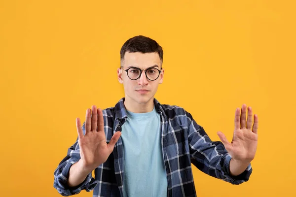 Mantén la distancia. Chico joven irritado en gafas que muestra el gesto STOP con ambas palmas en la cámara sobre fondo naranja — Foto de Stock