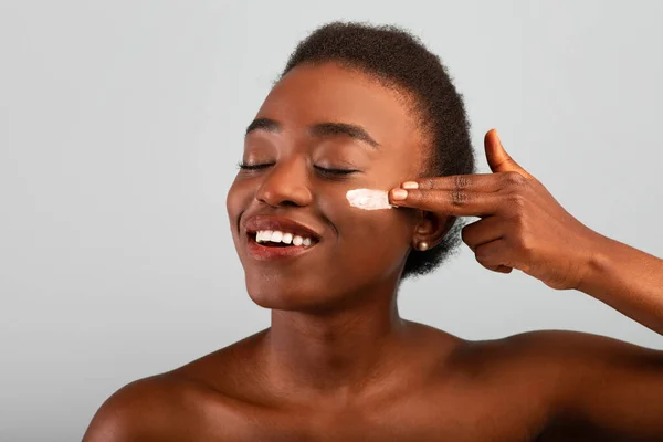 Codzienna pielęgnacja skóry. Afroamerykanka stosująca krem nawilżający na policzek, stojąca z zamkniętymi oczami na szarym tle — Zdjęcie stockowe