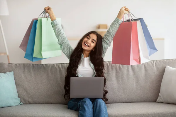 Emocionada señora india de compras en línea, sentado en el sofá con el ordenador portátil, levantando bolsas de regalo por encima de su cabeza en casa — Foto de Stock