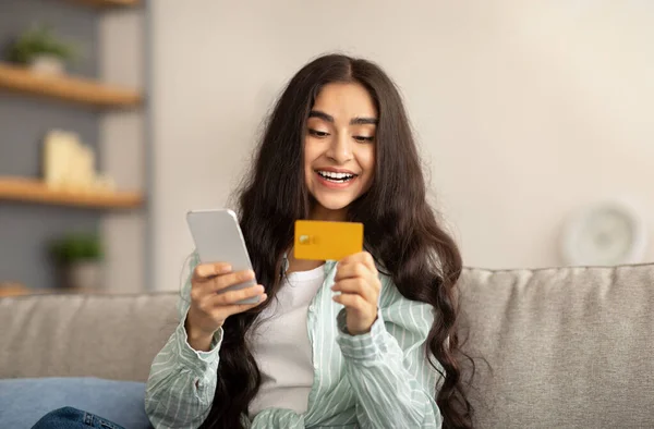 Portrait of cheerful Indian woman with mobile phone and credit card purchasing goods or services on web indoors — Stock Photo, Image