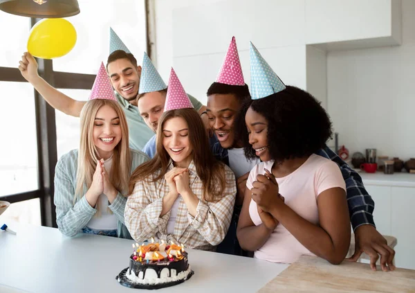 Pede um desejo. Amigos multirraciais alegres em bonés de festa comemorando juntos, soprando velas no bolo de aniversário em casa — Fotografia de Stock