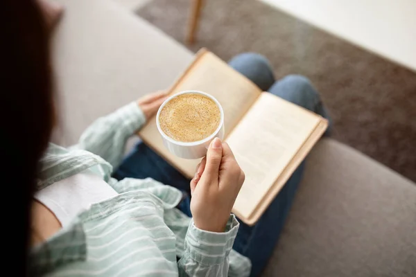 Relajante mañana de fin de semana. Vista recortada de la mujer india con taza de café y libro sentado en el sofá, primer plano — Foto de Stock