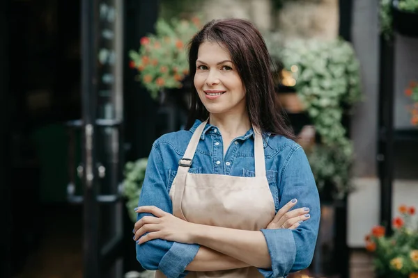 Усміхаючись молода приваблива впевнена жінка продавець в фартусі схрестив руки на грудях — стокове фото