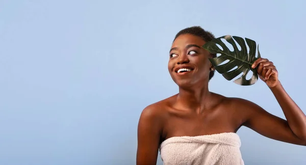 Africano americano mulher com pele lisa segurando folha monstera e olhando para o espaço livre no fundo estúdio azul — Fotografia de Stock