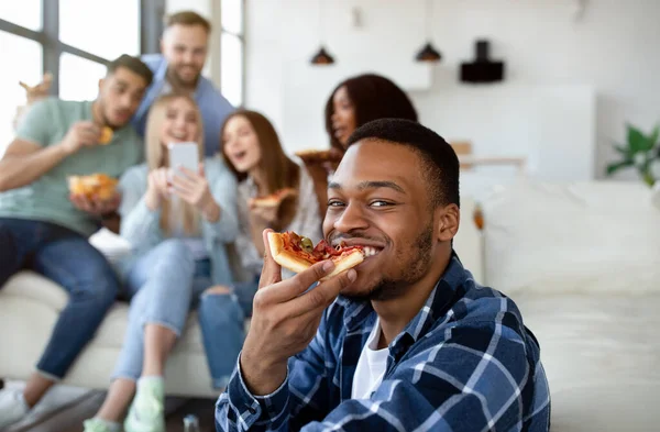 Knappe zwarte man eten plak pizza, groep van zijn multi-etnische vrienden met smartphone op de achtergrond — Stockfoto