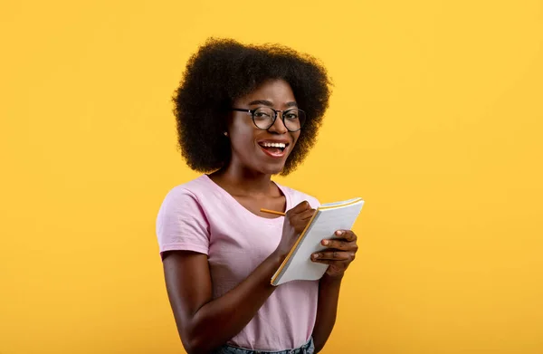 Checklist concept. Smart black female student wearing glasses, taking notes in copybook on yellow studio background — Stock Photo, Image