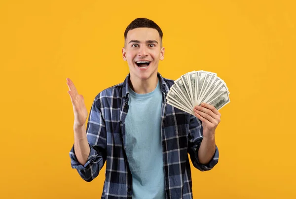 Joven guapo feliz por ganar una gran suma de dinero, sosteniendo billetes de dólar en fondo de estudio naranja —  Fotos de Stock