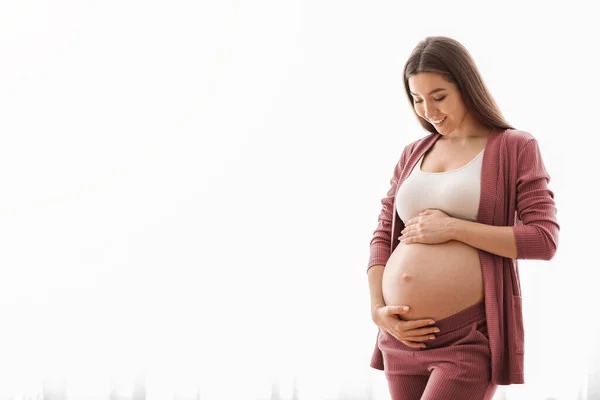 Mujer joven esperando bebé abrazando el vientre mientras está de pie cerca de la ventana en casa — Foto de Stock