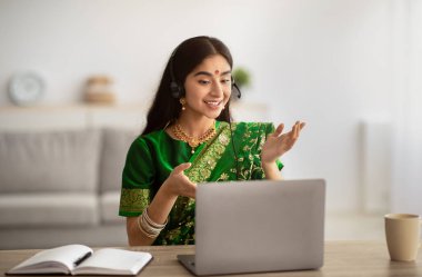 Positive Indian woman in traditional sari dress wearing headphones, having online meeting on laptop at home