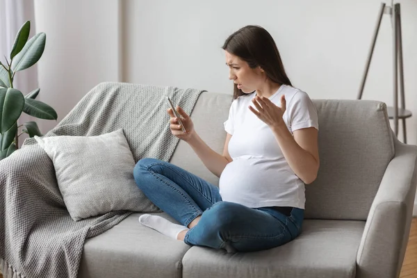 Zestresowana młoda kobieta w ciąży patrząc na ekran smartfona podczas relaksu na kanapie — Zdjęcie stockowe