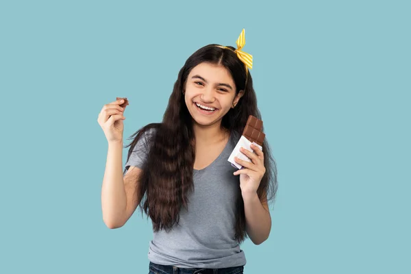 Portrait of happy Indian teen girl eating chocolate bar, feeling pleased on blue studio background — Stock fotografie