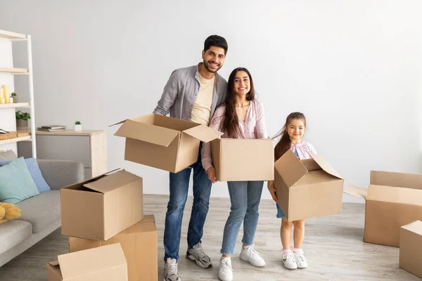 Щаслива східна сім'я тримає картонні коробки і посміхається на камеру, стоячи у своїй новій квартирі — стокове фото