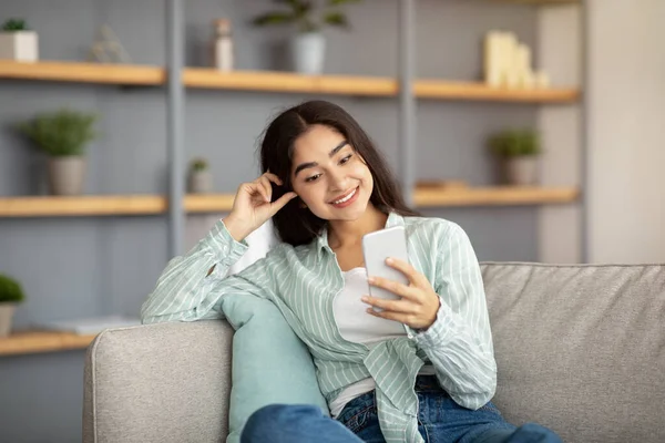 Portrait of happy Indian woman using smartphone on couch in living room, checking her email, posting in social media — Photo