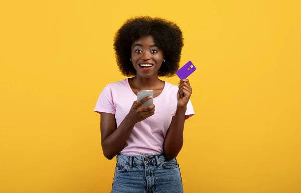 Easy money transfer. Positive black lady showing credit card and holding her mobile phone over yellow background — Stockfoto