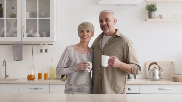 Felices cónyuges de mediana edad sosteniendo tazas de café posando de pie en la cocina — Vídeos de Stock