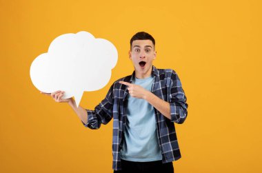 Cool young man pointing at empty speech bubble in excitement, orange studio background, mockup for design
