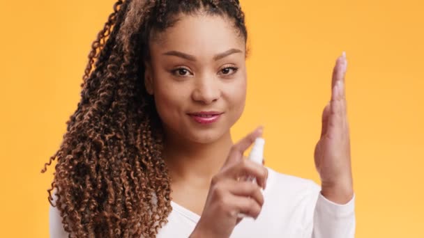 Joven mujer afroamericana positiva rociando desinfectante de alcohol líquido en la palma de la mano y sonriendo a la cámara, fondo naranja — Vídeo de stock