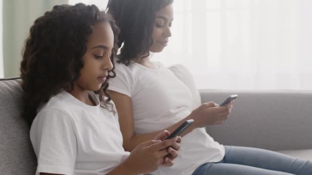작은 아프리카 계 미국인 여자 애와 스마트폰으로 그녀의 젊은 어머니 네트 워킹, 소파에 앉아서 서로를 무시 — 비디오