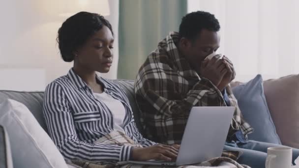 Africano senhora americana trabalhando distantemente em casa, seu marido doente beber chá médico, sentados juntos no sofá em casa — Vídeo de Stock