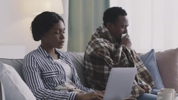 Young african american woman typing on laptop, her sick husband suffering from cough attack, sitting together at home — Stock Video