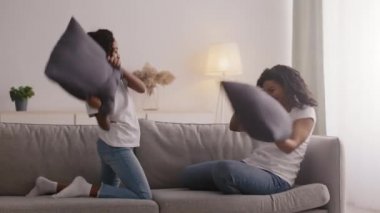 Mutlu Afro-Amerikan anne ve kızı yastık savaşı yapıyor, kanepeye düşüyor, sarılıyor ve birlikte gülüyorlar.