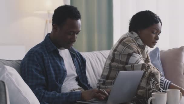 Młody czarny mężczyzna pracuje na laptopie w domu, jego chora żona kicha i dmucha w nos, siedzi owinięty w kratę na kanapie — Wideo stockowe