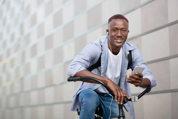 Chico negro guapo con mochila, auriculares y dispositivo móvil montar en bicicleta cerca de la pared de ladrillo en el centro, espacio en blanco — Foto de Stock