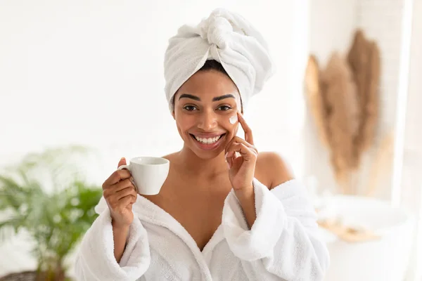Afrikaanse vrouw toepassen van crème op gezicht drinken koffie in de badkamer — Stockfoto
