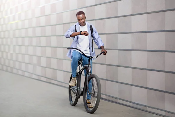 Feliz chico afroamericano con auriculares escuchando música y comprobando el tiempo en smartwatch durante el paseo en bicicleta fuera — Foto de Stock