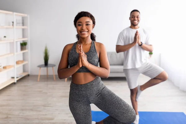 Neşeli zenci kadın erkek arkadaşıyla yoga yapıyor, ağaç duruşu yapıyor, namaste hareketi yapıyor, kapalı alanda. — Stok fotoğraf