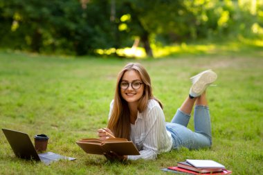 Öğrenme Konsepti. Gözlüklü Gülümseyen Öğrenci Kız Açık havada çalışırken kitap okuyor.