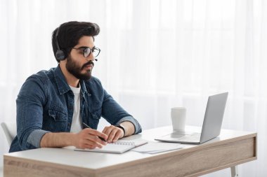 Çevrimiçi eğitim kavramı. Kulaklıklı bir Arap dizüstü bilgisayarda webinar izliyor ve not alıyor.