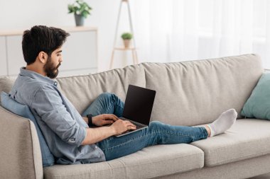 Rahat Arap adam boş ekranlı dizüstü bilgisayar kullanıyor, oturma odasındaki kanepede dinleniyor, iç mekan, boş alan, modelleme.