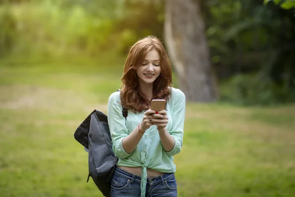 Chica alegre estudiante pelirroja utilizando el teléfono inteligente al aire libre, mensajes de texto con amigos — Foto de Stock