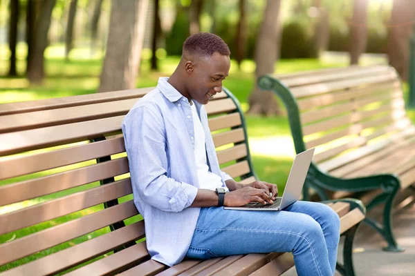 Πορτρέτο του δροσερό μαύρο άντρα χρησιμοποιώντας φορητό υπολογιστή στον πάγκο στο πάρκο, εργάζονται εξ αποστάσεως, επικοινωνώντας στην κάμερα — Φωτογραφία Αρχείου